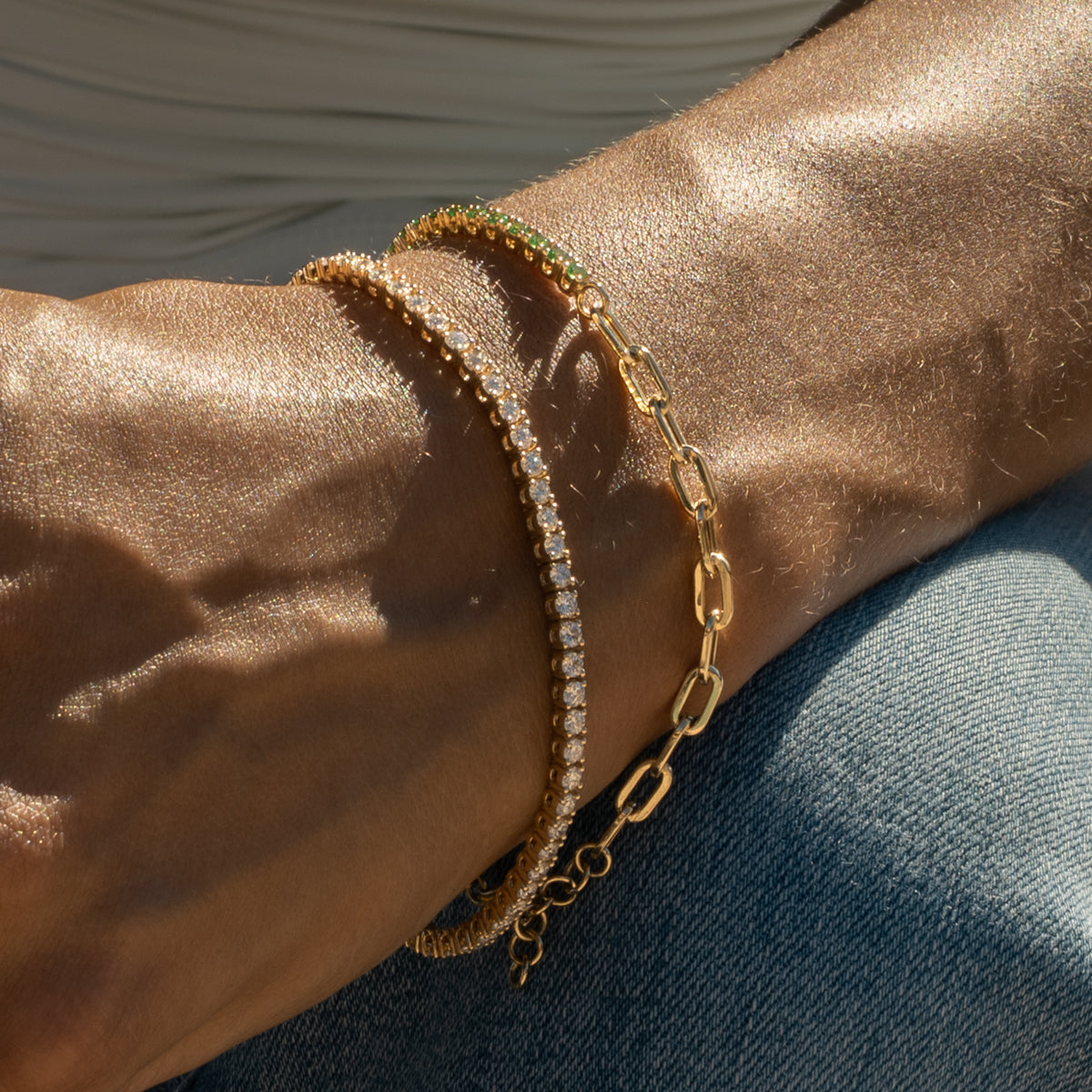 A close-up of a person's hand resting on blue denim jeans, wearing diamond and green garnet tennis bracelets on the wrist