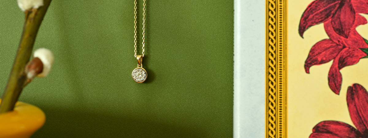 A gold pendant necklace with a round, clear-stone-set charm hangs against a dark green background, with decorative floral elements and a patterned frame partially visible at the edges.