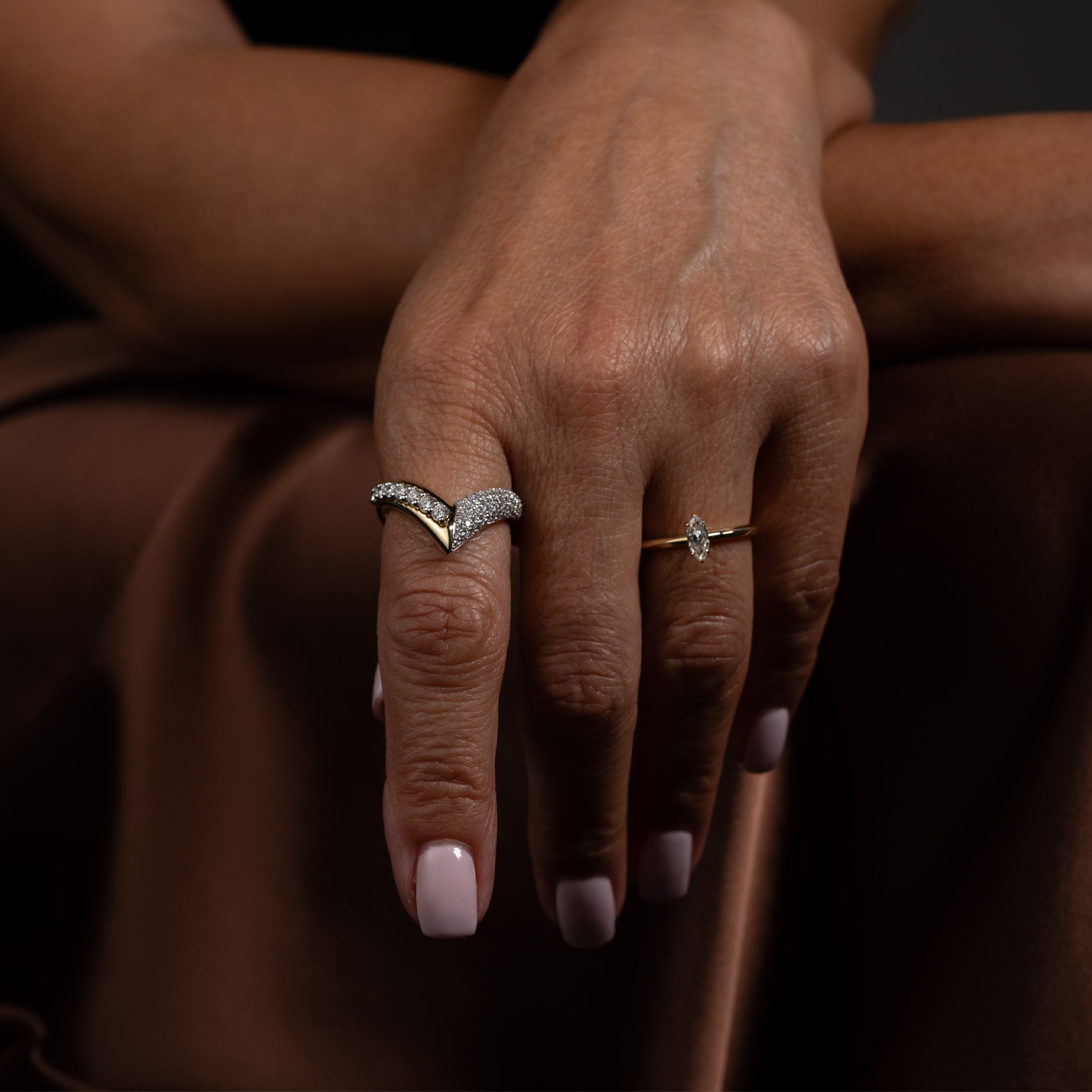 Close-up of a model wearing a bold gold and diamond ring paired with a marquise-cut solitaire ring against a neutral background.