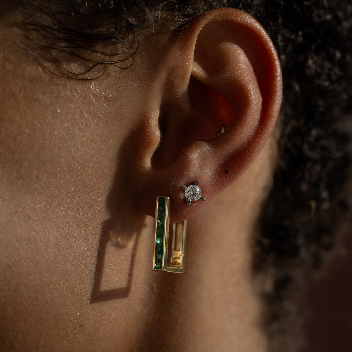 a close up to woman's ear, wearing an emerald earring matching with a classic four prong diamond stud