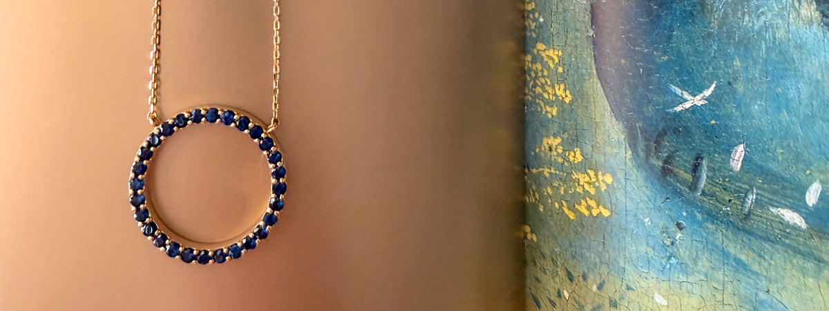 A gold circular pendant necklace with a border of deep blue sapphires, shown hanging against a warm beige background with a textured blue and yellow artwork partially visible on the right.