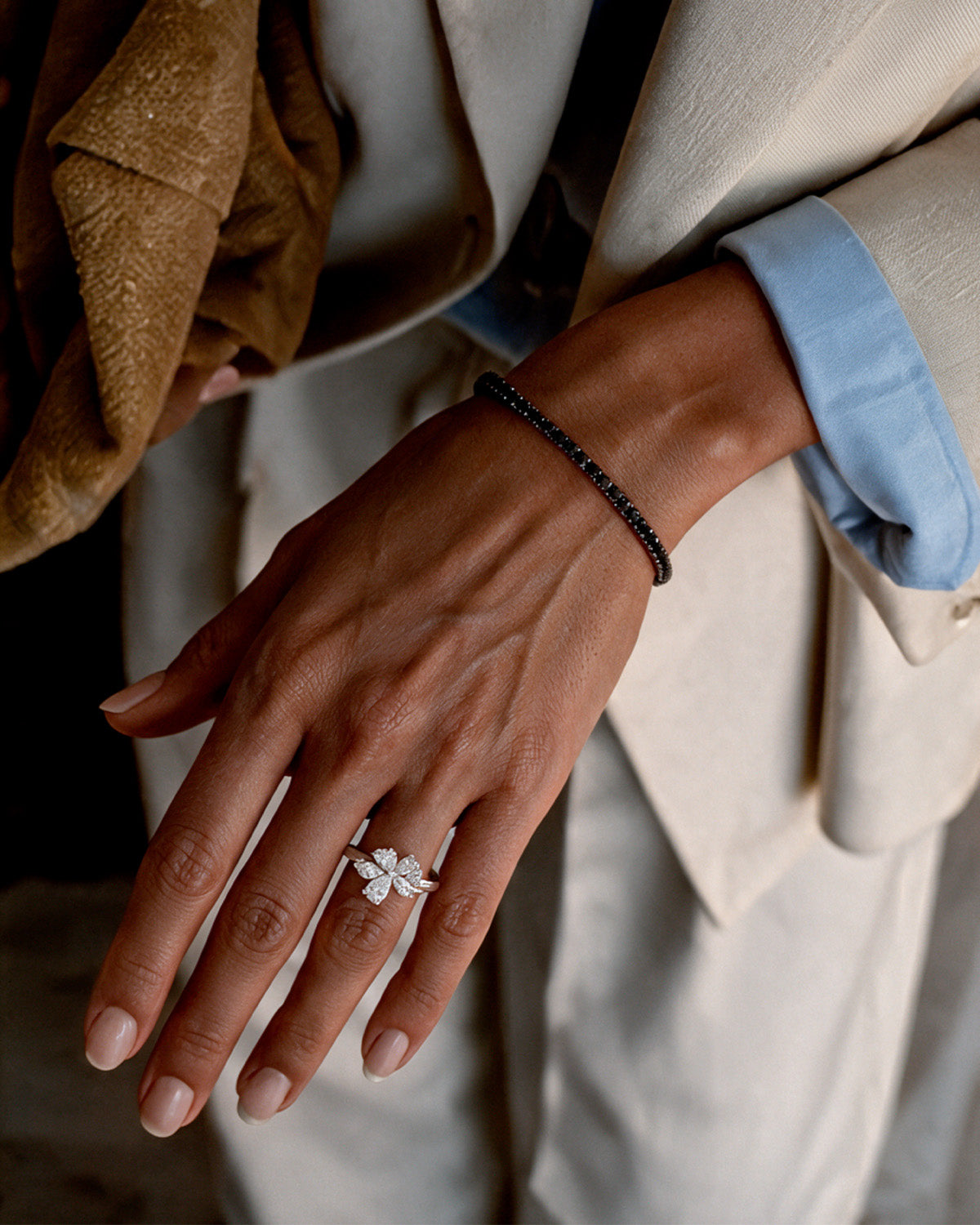 A close-up of a person's hand wearing a delicate black diamond bracelet and a white gold diamond ring with a white flower motif, styled with a light blue shirt and beige blazer.