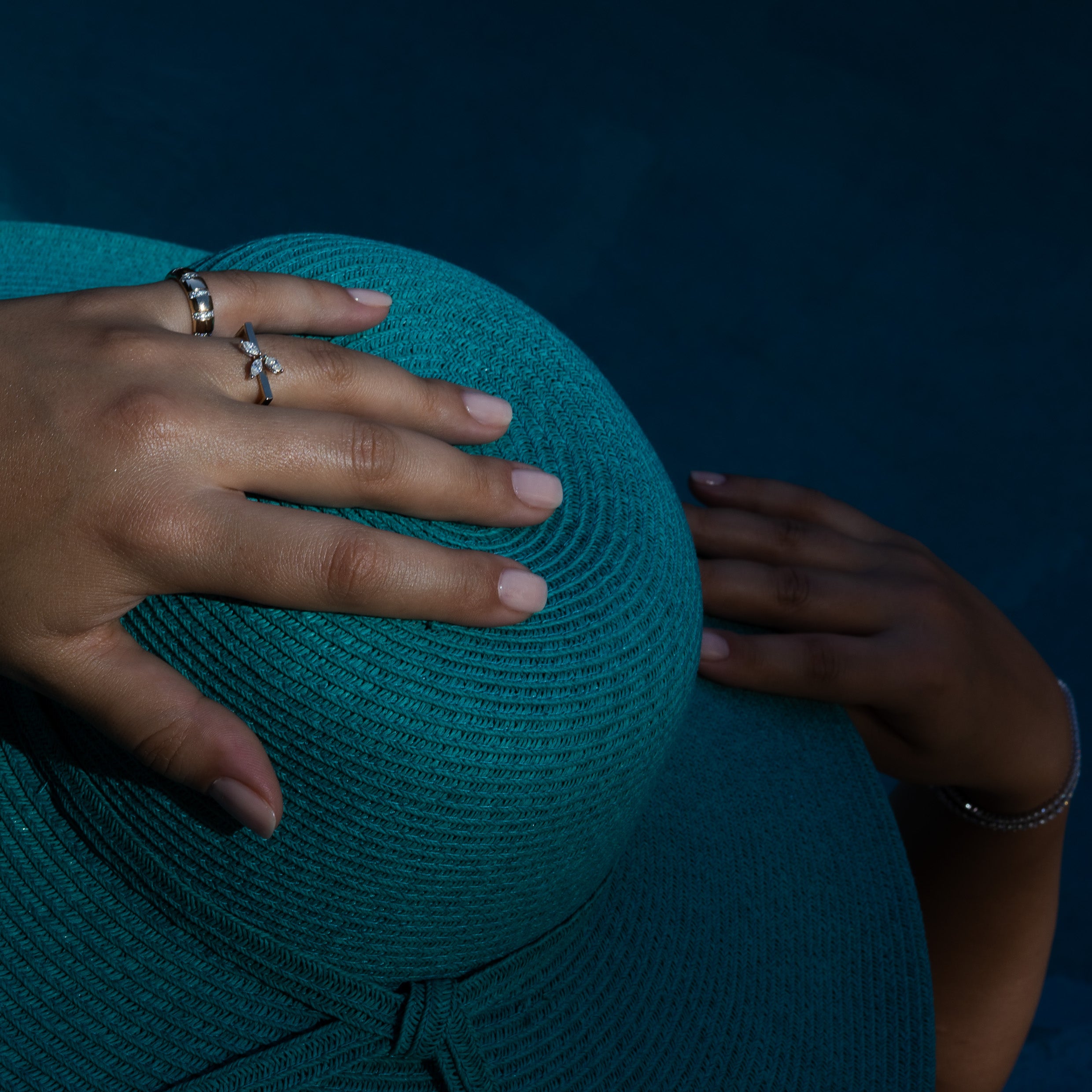 Teal straw hat with a hand wearing diamond rings holding it against a pool background