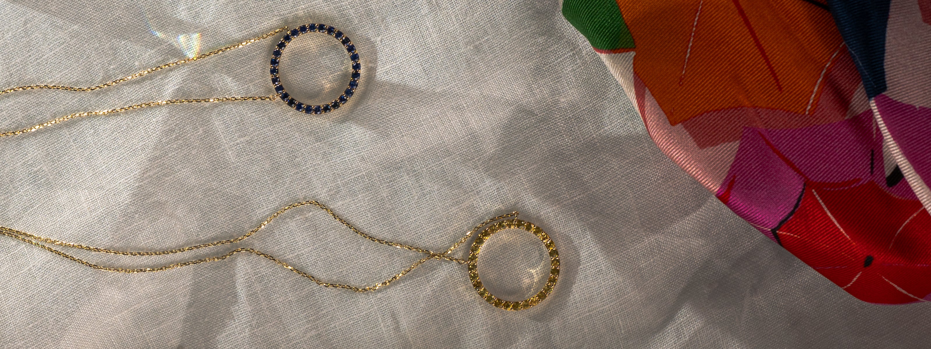 Two 14K gold necklaces featuring circular gemstone pendants resting on a textured surface, near a silk scarf.