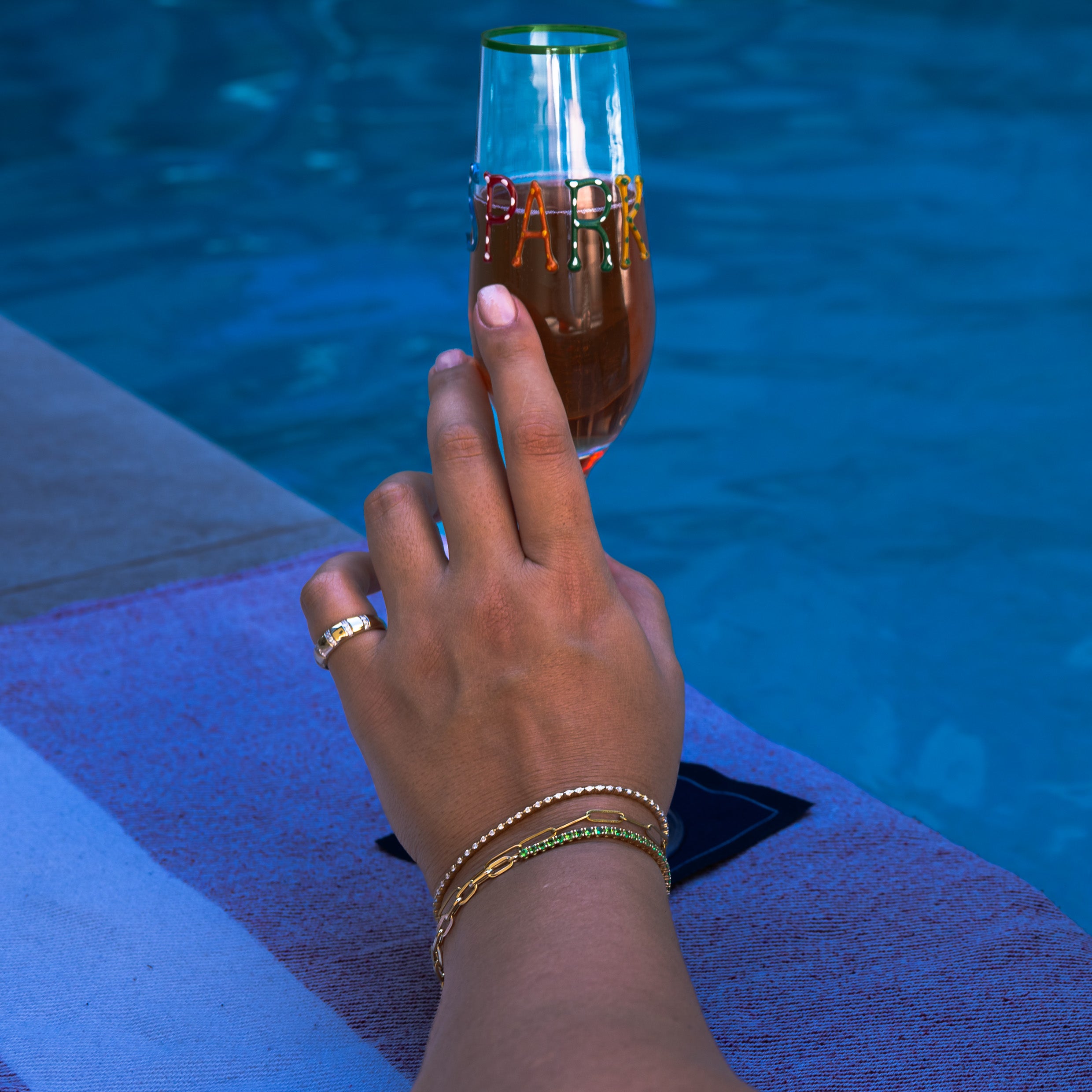 Hand wearing diamond tennis bracelets and holding a glass of champagne by a pool