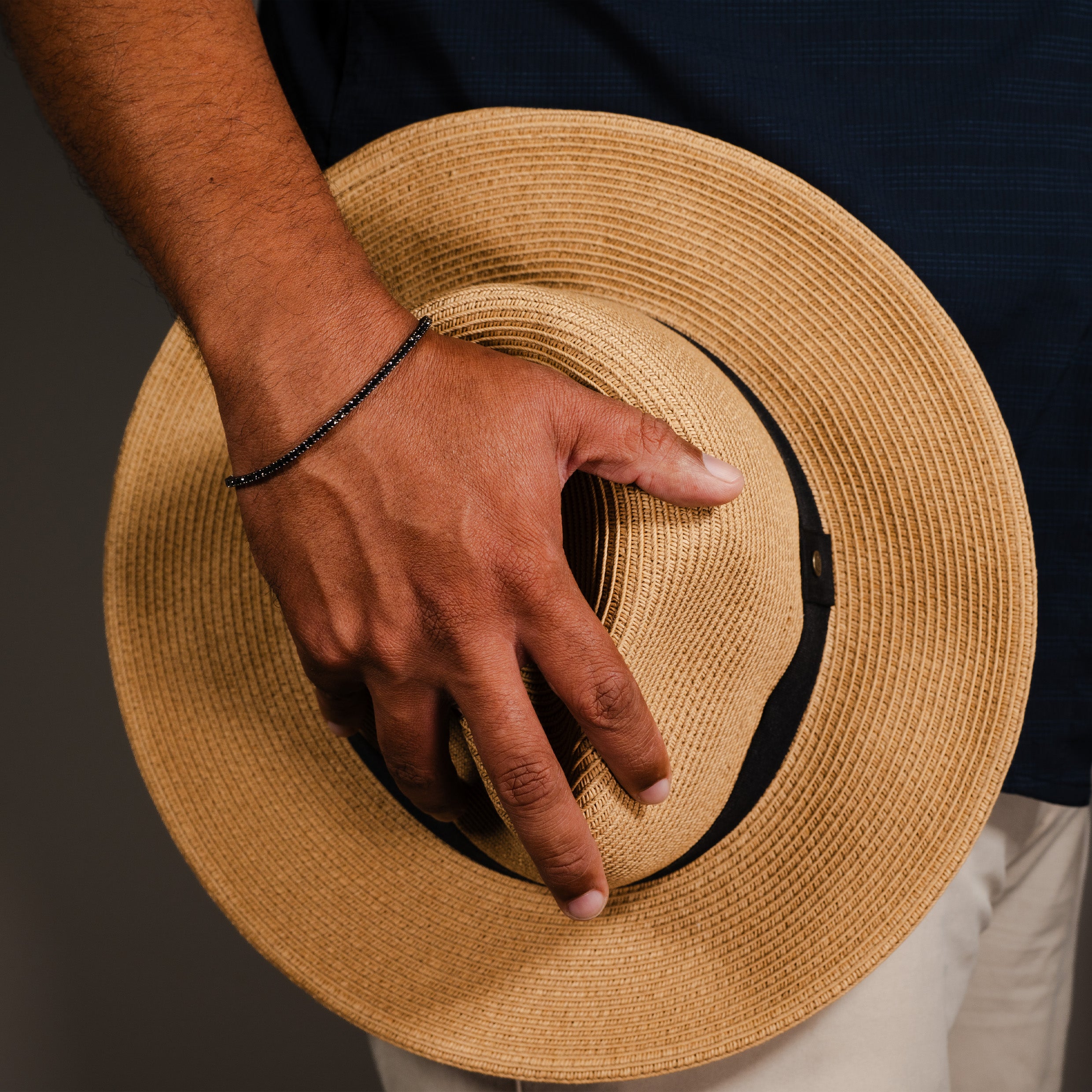 Man wearing a 14K gold black diamond tennis bracelet while holding a straw hat against a dark background.