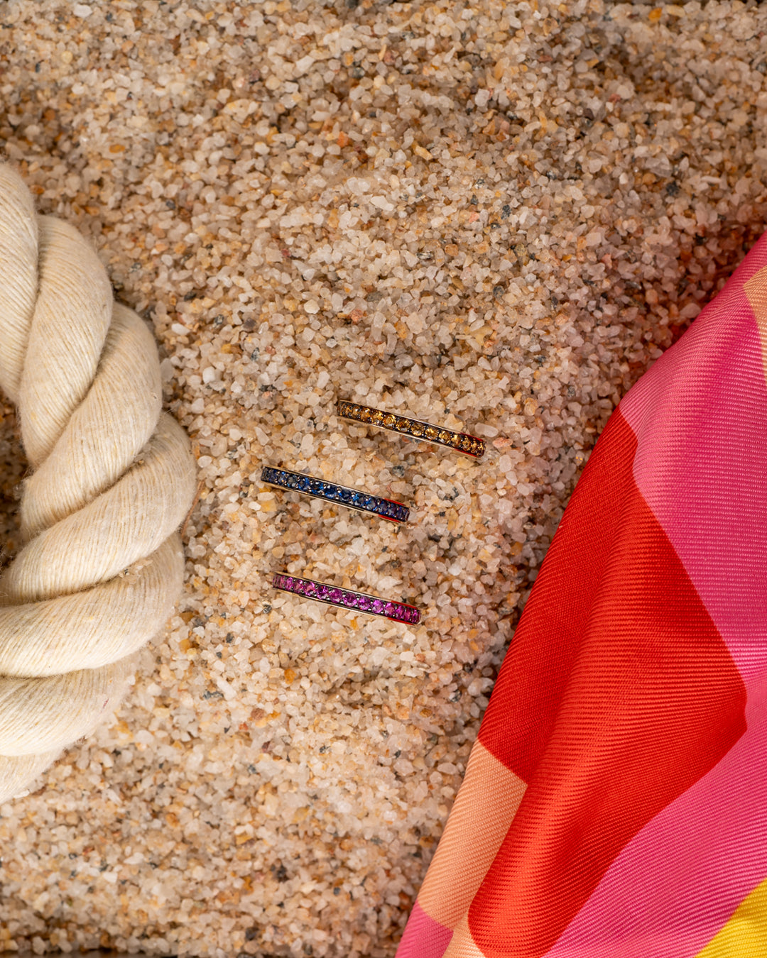 Three colorful sapphire rings on sand next to a silk scarf