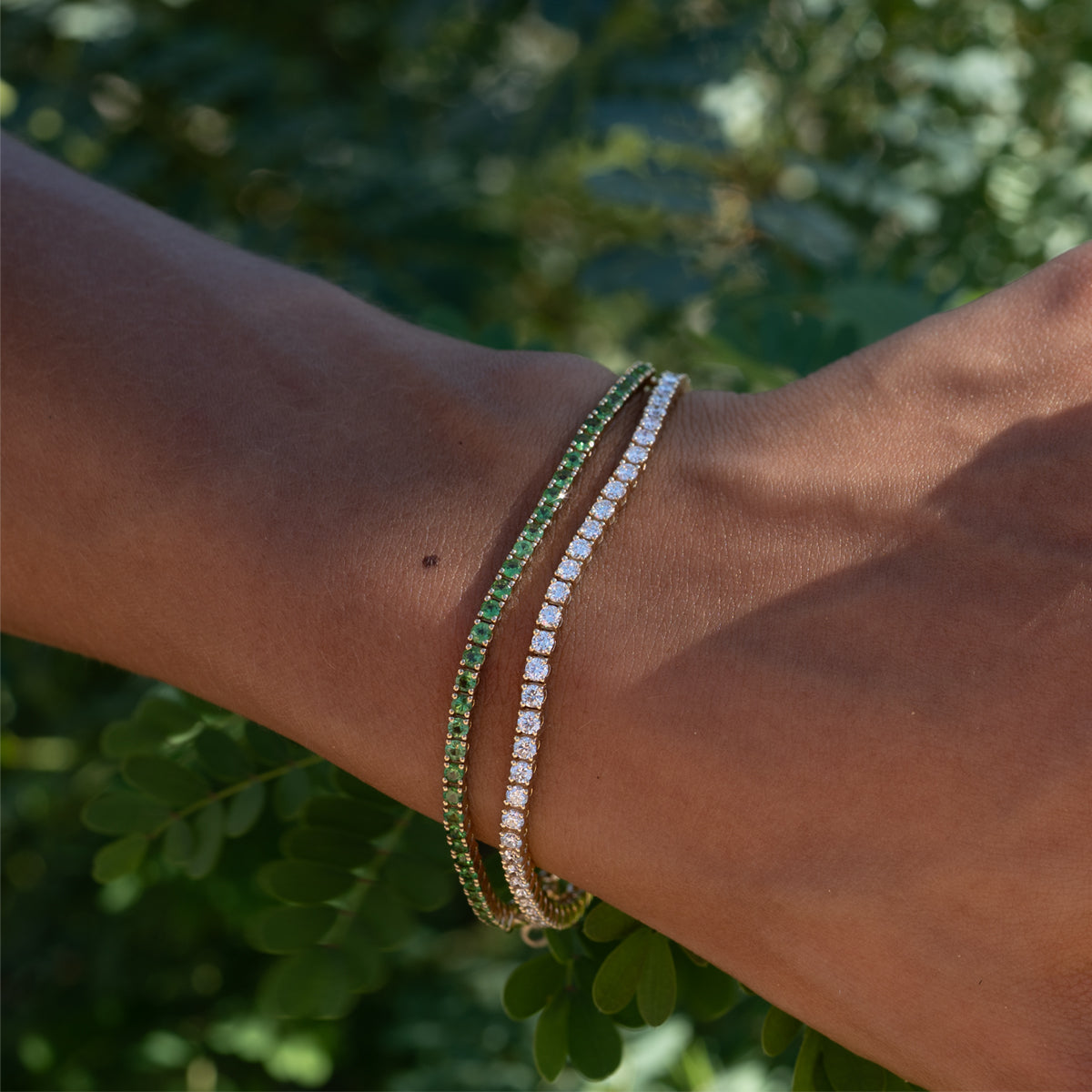 a close up photo of woman's wrist wearing diamond and green garnet tennis bracelets in a garden