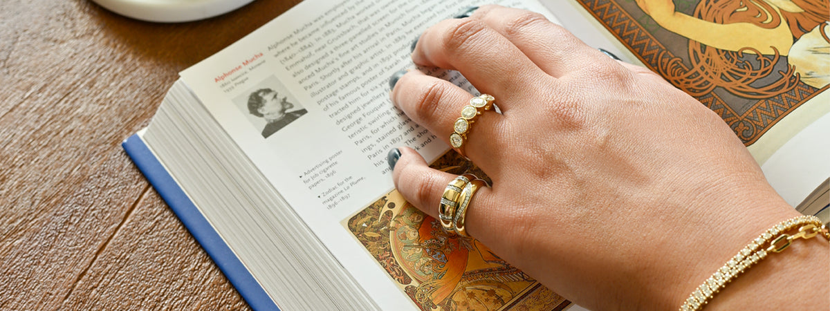 A close-up lifestyle shot of a hand resting on an open book, adorned with multiple gold rings and a gold bracelet.