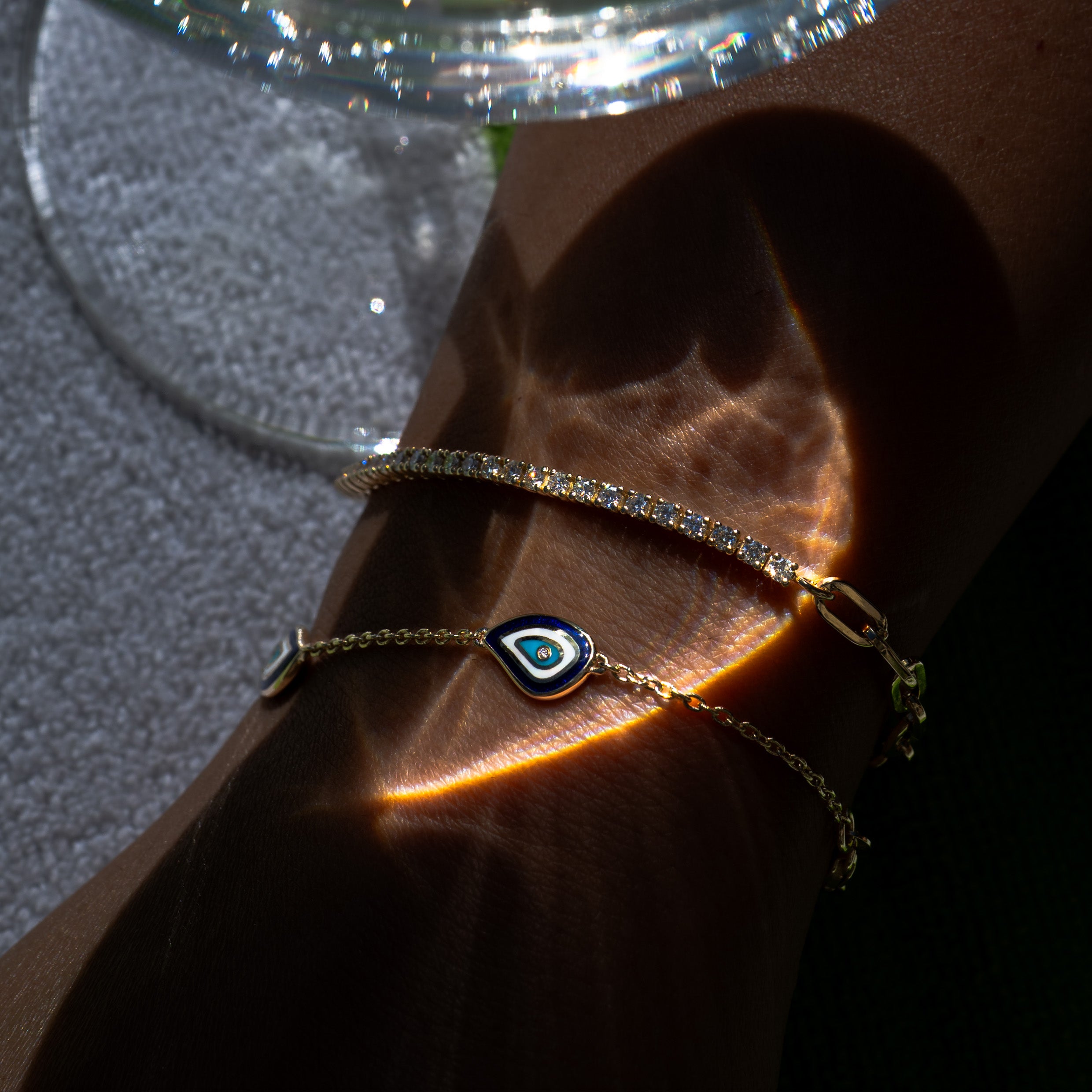 Solid gold diamond tennis bracelet and evil eye bracelet under dramatic light reflections