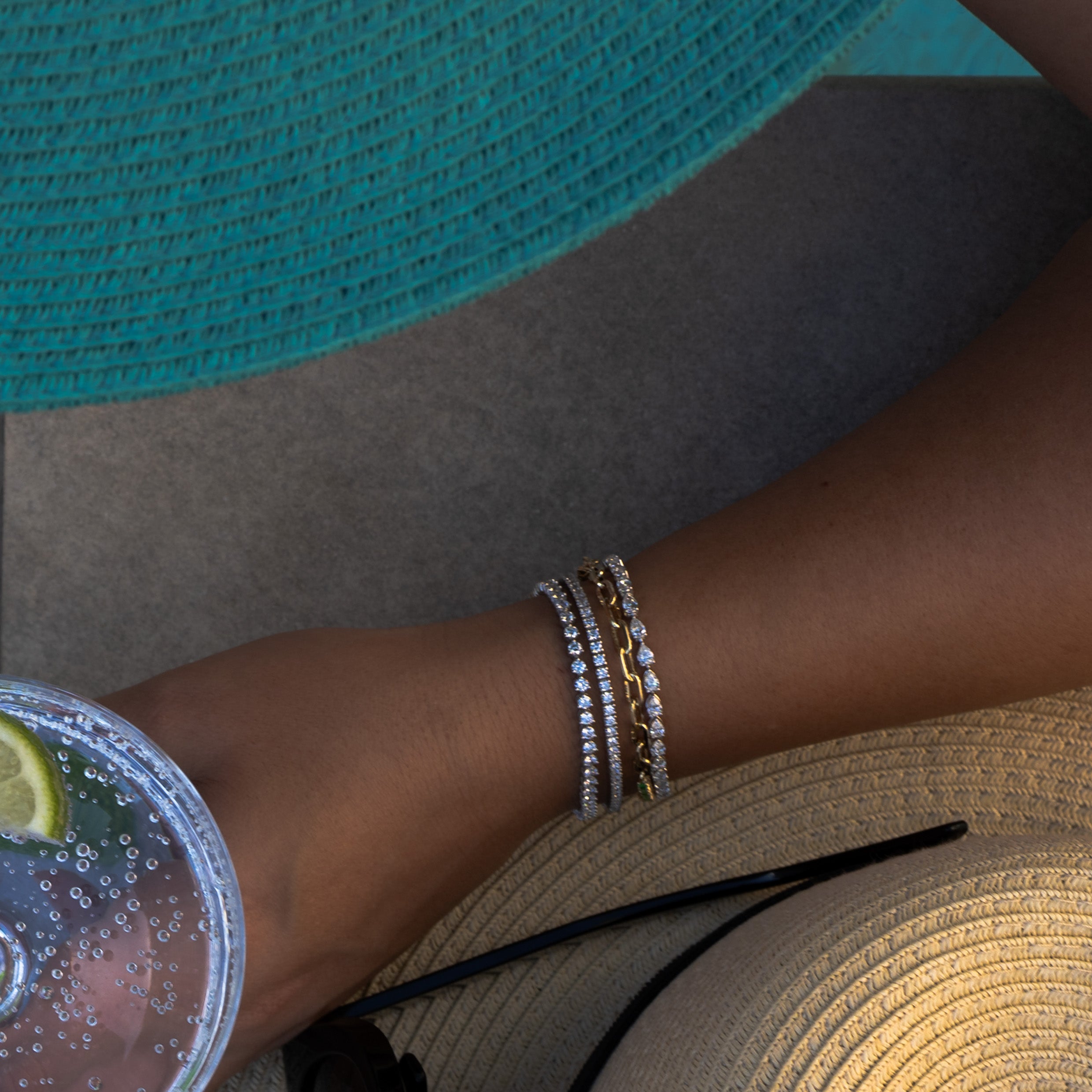 A woman's arm with diamond tennis bracelets holding a drink, wearing a teal hat.